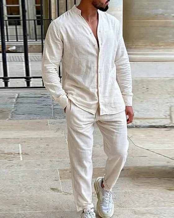 Ein Mann trägt ein naturweißes Leinen-Set, bestehend aus einem Langarmhemd mit Stehkragen und einer langen Hose, und steht in einem Innenhof mit Steinfliesen.
